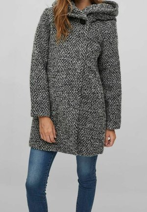 Kobieta w długim, szarym, teksturowanym, dzianinowym płaszczu z dużym kołnierzem i guzikiem, w połączeniu z niebieskimi dżinsami, stojąca na jednolitym tle.