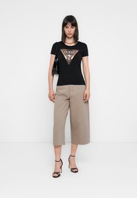 Sort kortærmet t-shirt med et trekantet "GUESS?" logo, parret med lyse beige bredbensbukser og sorte strophæle, mens der holdes en lille sort taske.