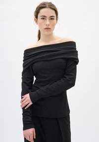 Mladá žena s hnědými vlasy v nízkém culíku má na sobě černý top s odkrytými rameny a dlouhými rukávy a černé kalhoty, stojí před bílým pozadím.