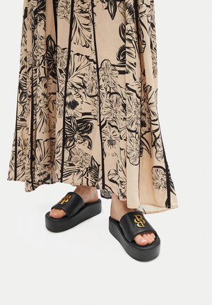 Čierne platformové sandále s hladkým povrchom, ktoré majú široký remienok a embosované zlaté logo. Na pozadí sa nachádza dlhá sukňa s kvetinovým vzorom.