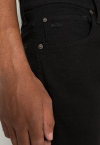 Černé džíny z denimu mají slim fit, decentní prošívání a kovové detaily. Materiál má hladkou texturu a mírný lesk.