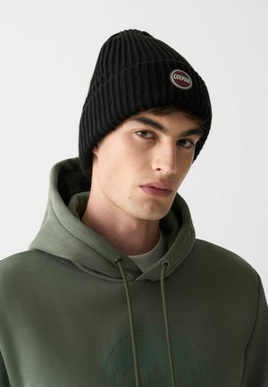 Cappellino beanie nero a costine con un badge rotondo con il logo Colmar, indossato sopra una felpa con cappuccio verde chiaro con una grande grafica circolare e coulisse.