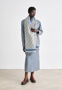 Gehaakte sjaal in het wit en blauw met geometrisch patroon, met franjes, gedragen over een spijkerjas en een blauwe rok, gecombineerd met bruine enkellaarsjes.