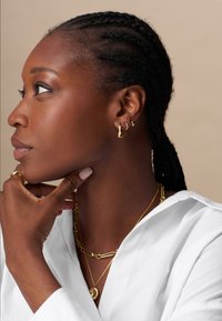 Žena v bielom topi je zdobená zlatými kruhovými náušnicami a vrstvenými náhrdelníkmi, pričom jej opletené vlasy rámujú profil na neutrálnom pozadí.