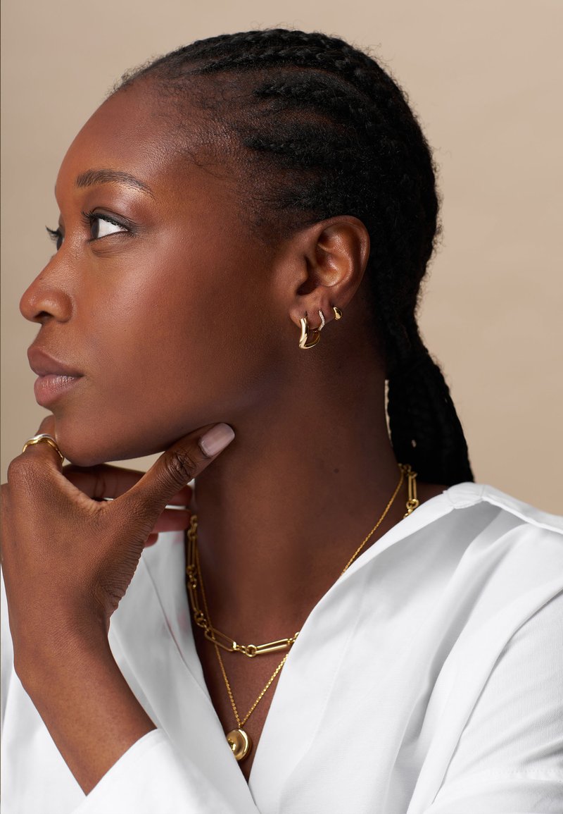 Žena v bielom topi je zdobená zlatými kruhovými náušnicami a vrstvenými náhrdelníkmi, pričom jej opletené vlasy rámujú profil na neutrálnom pozadí.