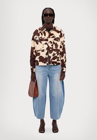 Cow print denimjacka med framfickor, åtföljd av ljusblå oversize jeans. Modellen håller en brun väska och bär mörka solglasögon.