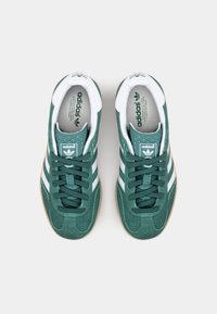 Gröna mockasneakers med vita ränder, en texturerad tunga och en gummisula. Designen inkluderar en rund tå och snörning fram.