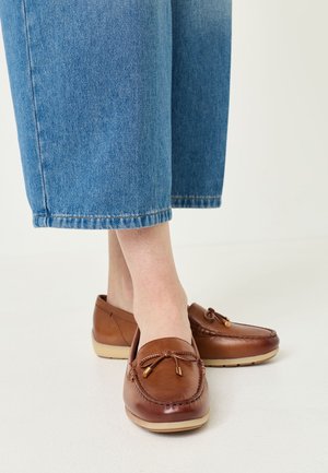 Bruine leren mocassin loafers met strikdetails gedragen met cropped blauwe spijkerbroeken op een lichte vloerachtergrond.