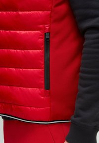 Rote Steppweste mit gequilteter Textur, seitlichem Reißverschluss und schwarzem dehnbarem Stoff an den Ärmeln und Seiten. Weiße Borte am Saum.