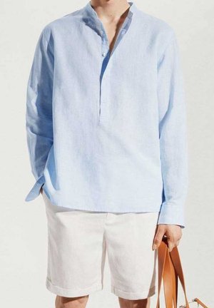Personne portant une chemise bleu clair à manches longues et un short blanc, tenant un sac en cuir marron à bandoulière dans une main sur un fond uni.