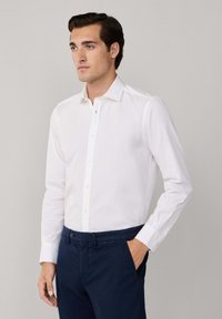 Hombre vestido con una camisa blanca de botones y pantalones azul marino, de pie con una mano en el bolsillo, mirando ligeramente hacia la izquierda contra un fondo simple.