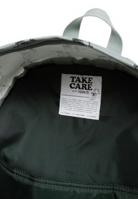 Grön ryggsäck inuti med en mjuk, mörk tygfoder och en etikett med texten "Var försiktig med nötter", samt tryckta tecknade figurer på innerlocket.