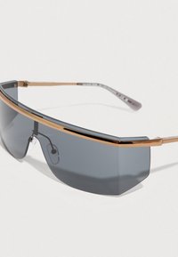 Κομψά γυαλιά ηλίου τύπου wraparound με σκούρο φακό σε σχήμα ασπίδας και λεπτό χρυσό πλαίσιο σε λευκό φόντο.