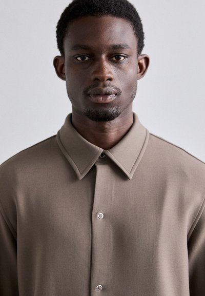 Ung mand med kort krøllet hår iført en taupe knap-shirt, som står med ansigtet fremad mod en ensfarvet lys baggrund.