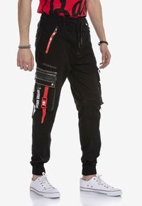 Schwarze Cargo-Hosen mit mehreren Taschen, Reißverschlüssen und roten Akzenten. Elastische Bündchen und ein Kordelzug am Bund vervollständigen das utilitaristische Design.