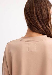 Sudadera beige claro con cuello redondo y un detalle de parche cosido en la parte posterior. La tela parece suave con una textura blanda.