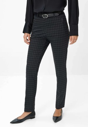 Woman wearing black houndstooth slim-fit pants, black belt, black long-sleeve blouse, and black pointed flats, standing against a plain background.