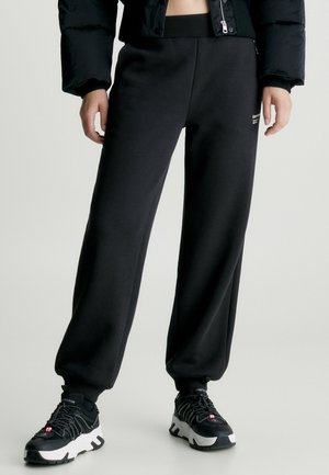 Personne portant un pantalon de survêtement noir avec des poignets élastiques, des baskets noires et blanches, et une veste courte noire sur un fond uni.