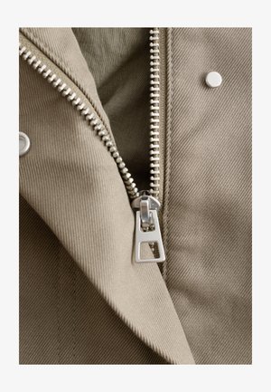 Nahaufnahme eines teilweise geöffneten silbernen Reißverschlusses an einem beigen Kleidungsstück mit sichtbarem Druckknopf und Nahtdetails.