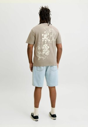 Mann med dreadlocks står med ryggen til iført beige t-skjorte med hvit koi-fiskdesign og lyseblå denimshorts med svarte joggesko.