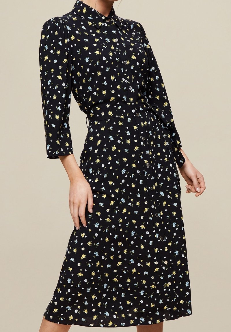 Abito floreale nero con motivi gialli e blu, design con colletto, maniche lunghe, lunghezza media, tessuto leggero e chiusura con bottoni.