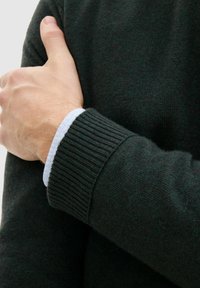 Dunkelgrüner Strickpullover mit gerippten Bündchen, über einem hellblauen Hemd getragen. Weiche Textur, die die feine Detail- und Qualitätsstruktur des Stoffs zeigt.