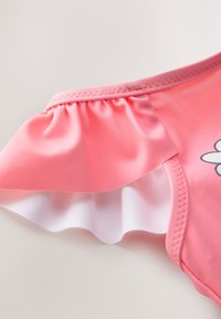 Rožinės spalvos maudymosi kostiumėlis su rauktu petnešėliu. Turi minkštą tekstūrą ir baltą uždaigą po raukiniu. Ant priekio gėlių raštas.