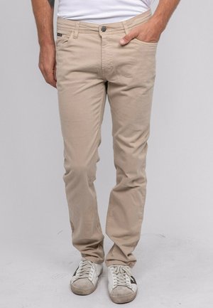 Homme portant un pantalon beige décontracté, des baskets blanches avec des rayures noires, debout avec une main dans la poche sur un fond uni.