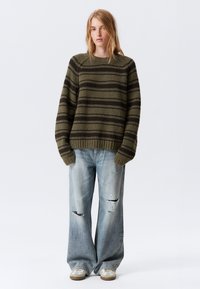 Strikkegenser i olivengrønn med mørke striper, med rund hals og ribbestrikkede kanter, kombinert med slitte lys azul brede jeans.