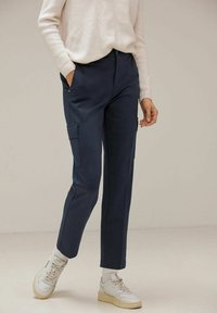 Pantalons cargo bleu marine avec poches latérales, design ajusté et poignets. Portés avec un pull en tricot crème et des baskets blanches. Texture lisse.