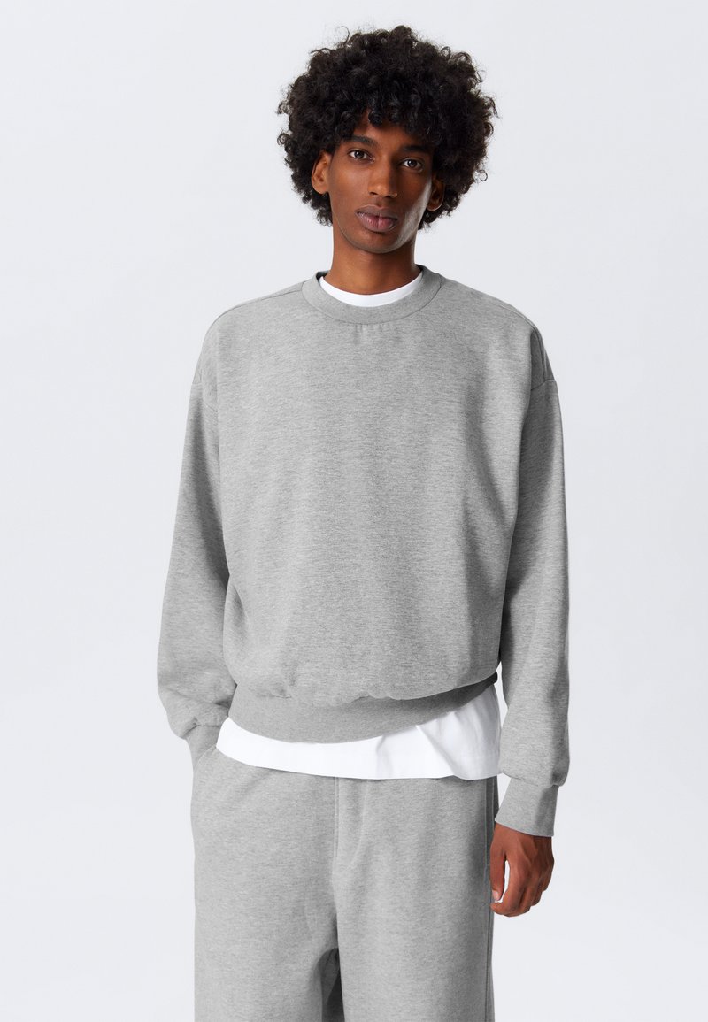 Grijze sweatshirt met lange mouwen, ronde hals en geribbelde boorden. Draagt over een wit shirt, wat een casual en ontspannen uitstraling geeft.