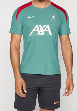Uomo che indossa una maglietta da allenamento del Liverpool FC di colore teal con accenti rossi sulle spalle e pantaloncini neri Nike con logo arancione, in piedi davanti a uno sfondo neutro.