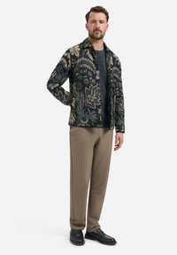 Man in een bloemenpatroon blousejack over een donker shirt, beige broek en zwarte nette schoenen, staand met een hand in zijn zak.