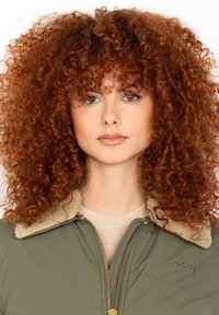 Donna con voluminose capelli ricci rossi, che indossa una giacca verde con un colletto in pelliccia sintetica beige, di fronte con un'espressione neutra.