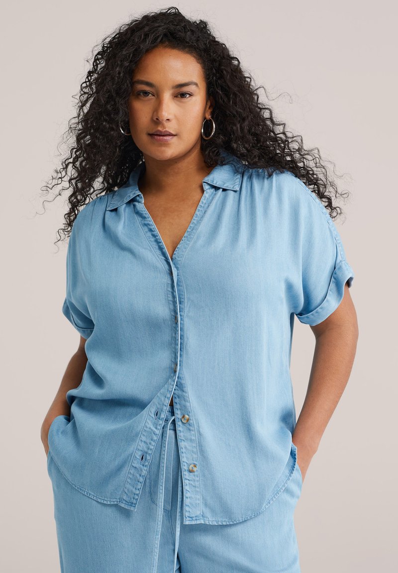 Naine pikkade lokkis juustega, kannab helesinist lühikese varrukaga nööpidega särki ja sobivaid pükse, käed taskutes, neutraalne taust.