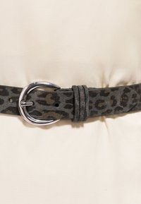 Curea gri din piele intoarsă cu pete negre de leopard și o cataramă metalică argintie strălucitoare fixată în jurul unui material bej deschis.