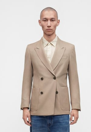 Homme à la tête rasée portant un blazer beige croisé sur une chemise beige clair et un jean bleu, debout devant un fond blanc uni.