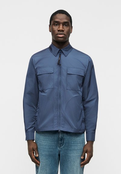 Jeune homme portant une veste zippée bleue avec des poches sur la poitrine et un col pointu, associée à un jean bleu clair sur un fond uni.