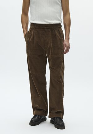 Mees, kes kannab pruune sametpükse, musti kingi ja valget äravarrukateta soonikutest särki, seisab käed taskutes tavalise tausta ees.