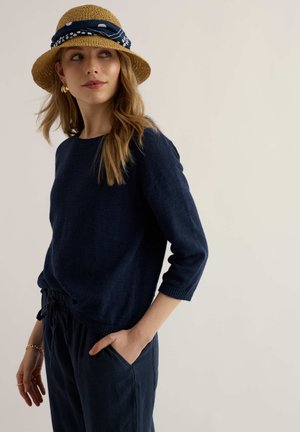 Donna con maglione e pantaloni blu navy e cappello di paglia decorato con una sciarpa blu navy con motivo, che guarda di lato con una mano in tasca.