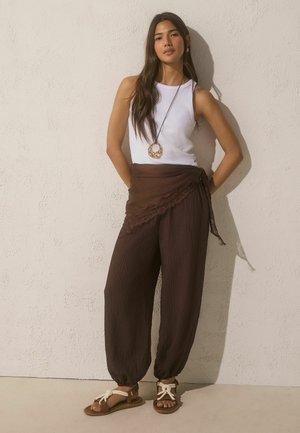 Junge Frau steht vor einer beigen Wand, trägt ein weißes ärmelloses Oberteil, braune lockere Hosen mit Wickelfunktion, Sandalen und eine lange Halskette mit Anhänger.