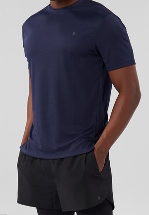 Homme portant un t-shirt de sport bleu marine à manches courtes et un short de course noir, les mains dans les poches, sur un fond uni.