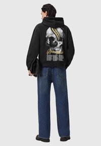 Un hoodie noir avec un graphic de crâne, un serpent et des accents floraux au dos ; un jean en denim bleu à coupe ample ; une personne tenant une pochette noire texturée.