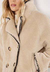 Beige fleece coat met een dubbelbreasted ontwerp, zwarte stiksels langs de randen, grote knopen en een hoge kraag voor extra warmte.