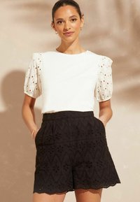 Bela bluzica z zanko in čipkastimi rokavi ter črne vezenine kratke hlače z valovitim robom, oblikovane z cvetličnimi izrezi.