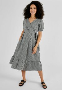 Rochie de gingham alb-negru cu decolteu în V, mâneci bufante, talie strânsă și tiv volanat. Modelul poartă sandale negre cu talpă plată, pozează cu mâinile în buzunare.