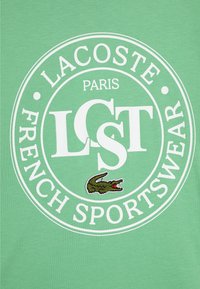 Grønn bomull t-skjorte med en rund hvit logo med "LACOSTE," "PARIS," og "FRENCH SPORTSWEAR," i tillegg til en brodert krokodilledetalj.