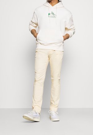 Person som bär en krämfärgad hoodie med en grön sneaker-grafik, krämfärgade byxor och vita sneakers, står med händerna i hoodie-fickorna.