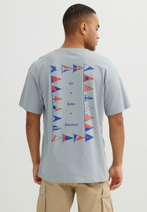 Hombre vistiendo una camiseta gris clara con banderas náuticas rojas y azules y texto "Les Belles Bannières" en la espalda, pantalones cortos cargo beige.