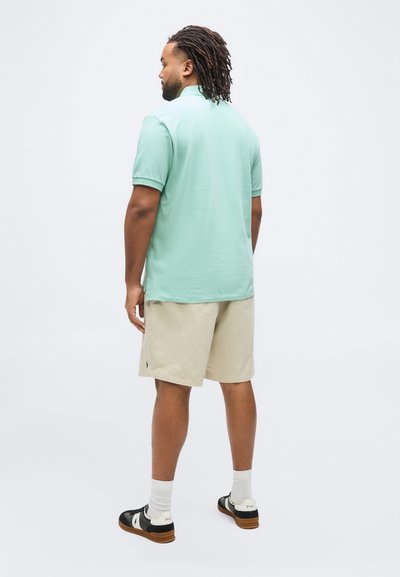 Άνδρας με dreadlocks που φοράει πουκάμισο πόλο μέντας, μπεζ σορτς, λευκές κάλτσες και ασπρόμαυρα αθλητικά παπούτσια, στέκεται γυρισμένος προς τα πίσω.